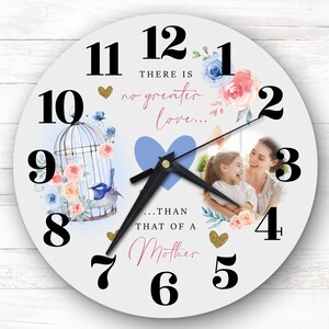 May include: A white clock with black numbers and hands. The clock has a floral design with a blue bird in a cage. The clock also has a photo of a mother and daughter with the text "There is no greater love... than that of a Mother".