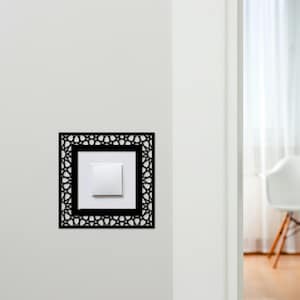 May include: A black, geometric patterned light switch cover is mounted on a white wall. The cover surrounds a white light switch. The design features a square frame with an intricate, openwork pattern. The background is a white wall and a glimpse of a room.