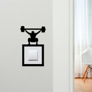 May include: A black silhouette of a weightlifter lifting a barbell, positioned above a light switch. The light switch is white, set within a black frame. The design is mounted on a white wall.