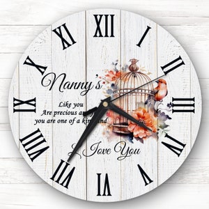 May include: A white wooden clock with Roman numerals and a black clock hand. The clock face features a watercolor illustration of a bird in a cage with flowers. The text "Nanny's" and "Like you are precious and you are one of a kind" is written on the clock face. The text "Love You" is written below the illustration.