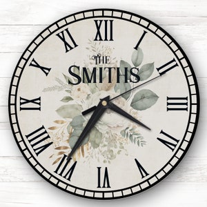 May include: A round clock face with a white and cream floral design featuring gold accents. The clock face has Roman numeral markings and a black clock hand. The text "The Smiths" is printed on the clock face.