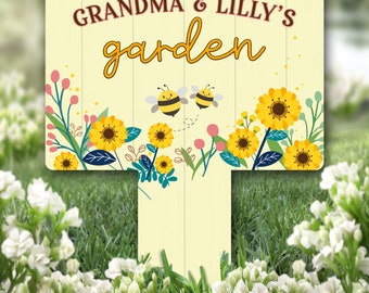 Può includere: Un cartello da giardino giallo con il testo "Grandma & Lilly's garden" in lettere marroni. Il cartello è decorato con girasoli gialli, foglie verdi e due api di cartone animato.