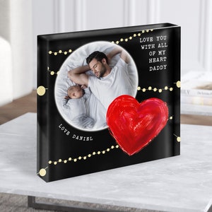 May include: A black square photo block with a white circle containing a photo of a father and baby sleeping. The photo is surrounded by a string of gold lights. A red heart with a painted texture is in the lower right corner. The text "Love you with all of my heart Daddy" is in the upper right corner. The text "Love Daniel" is in the lower left corner.