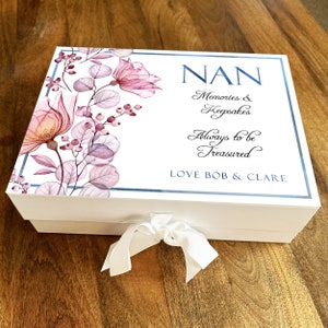 May include: White keepsake box with a floral design and blue trim. The box has a white ribbon and the text "NAN Memories & Keepsakes Always to be Treasured LOVE BOB & CLARE" on the lid.