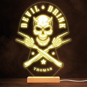 May include: A yellow neon sign with a skull and crossbones design. The skull has horns and a devilish grin. The text "DEVIL DRINK" is written in a circle around the skull. The text "THOMAS" is written below the skull and crossbones.