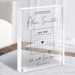 Peut inclure: Un bloc acrylique transparent avec le texte "OUR PRECIOUS BABY" et "Alex Smith" en écriture cursive noire. Un calendrier d'avril 2022 avec un cœur noir sur le 18. Le texte "We will Love You forever x" est écrit en écriture cursive noire.