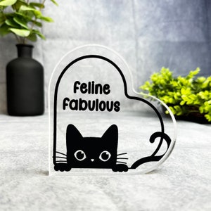May include: A clear heart-shaped acrylic sign with the text "Feline Fabulous" and a black silhouette of a cat peeking out from behind the text.