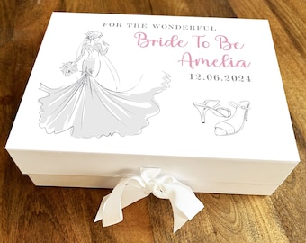 Puede incluir: Caja de regalo blanca con una cinta de satén blanca. La caja tiene un dibujo de una novia con un vestido de novia y un par de tacones altos. El texto en la caja dice "Para la maravillosa novia Amelia 12.06.2024".