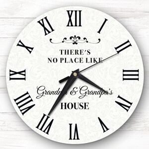 May include: White round clock with black Roman numeral hour markers and black hands. The clock face has a white background with a subtle floral pattern. The text "There's no place like Grandma & Grandpa's House" is printed on the clock face.