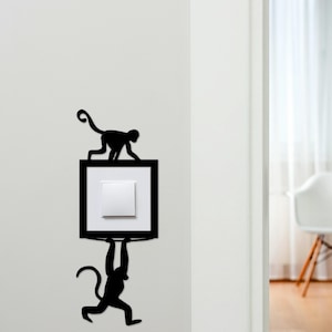 May include: Black metal monkey wall art. The design features two monkey silhouettes, one above and one below a light switch, adding a fun touch to any room. The light switch is white, set in a black frame.