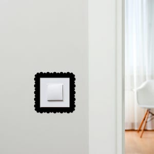 May include: A white light switch with a square, white faceplate is centered within a black, scalloped-edge frame. The frame is mounted on a plain white wall. The image is well-lit, with a minimalist aesthetic.