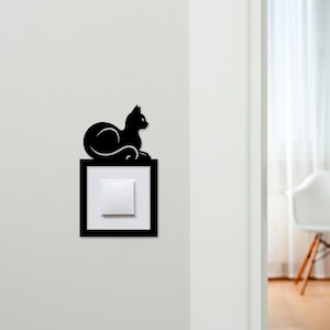 May include: A black cat silhouette decorates a light switch cover. The cat is in a relaxed pose, atop a black frame surrounding a white switch. The design adds a whimsical touch to a functional item. The wall is painted white.