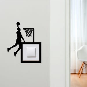 May include: Black silhouette of a basketball player mid-air, about to dunk the ball. The design is mounted on a wall, above a light switch. The basketball hoop and net are also black, creating a striking contrast against the white wall.