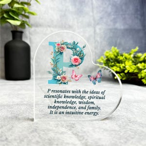 May include: A clear heart-shaped acrylic ornament featuring a blue letter P adorned with pink and white flowers. The ornament includes a quote about the letter P and its significance.