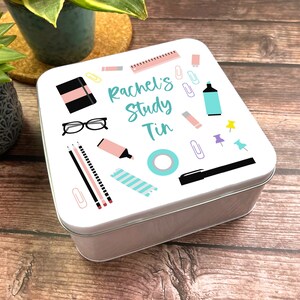 Puede incluir: Una lata cuadrada blanca con un diseño colorido de útiles escolares. El texto "Rachel's Study Tin" está impreso en la lata.