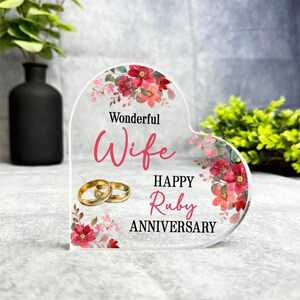 Könnte beinhalten: Eine transparente, herzförmige Acryl-Plakette mit einem Blumendesign und dem Text "Wonderful Wife Happy Ruby Anniversary". Zwei goldene Ringe sind auf der Plakette abgebildet.