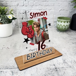 Puede incluir: Un marco de fotos de acrílico transparente con una base de madera. El marco presenta una foto de un joven, guantes de boxeo rojos, estrellas negras y el texto "Simon", "Feliz cumpleaños número 16".