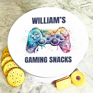 Op de afbeelding: Een witte ronde blik met een kleurrijk aquarelontwerp van een videogamecontroller. De tekst "WILLIAM'S GAMING SNACKS" staat op de blik gedrukt.