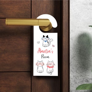 以下が含まれることがあります： 眼鏡と青いバンダナをつけた猫が描かれた白いドアハンガー。ピンク色の文字で「Amelia's Room」と書かれています。ハンガーの下には眼鏡をつけた猫が2匹います。