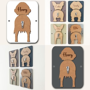 Op de afbeelding: Set van zes houten haken in de vorm van een hond met gepersonaliseerde namen. De haken zijn in verschillende kleuren, waaronder wit, grijs, lichtgroen en bruin. Elke haak heeft een metalen haak om spullen aan op te hangen.