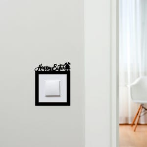 May include: A black decorative light switch cover with the words "Happy Easter" and a bunny silhouette. The cover is mounted on a white wall, framing a white light switch.