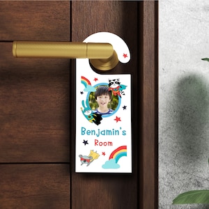 May include: A white door hanger featuring a rainbow, stars, and a photo of a child. The text on the door hanger reads "Benjamin's Room".