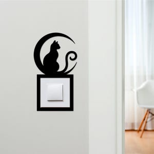 May include: Black cat silhouette on a crescent moon above a light switch. The cat's tail curls. The light switch is framed in a black square. The design is mounted on a white wall. The overall design is a decorative wall feature.