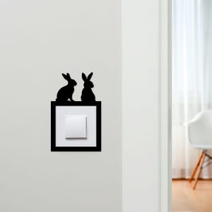 May include: Black silhouette of two rabbits perched above a white light switch. The light switch is set in a black square frame, mounted on a white wall. The rabbits are facing opposite directions.