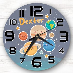 May include: A round wall clock with a gray background featuring cartoon planets and stars. The clock face has large black numbers and the name "Dexter" in orange. The clock hands are black.