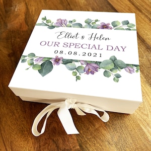 May include: White gift box with a white ribbon. The box has a floral design with purple flowers and green leaves. The text on the box reads "Elliot & Helen Our Special Day 08.08.2021".