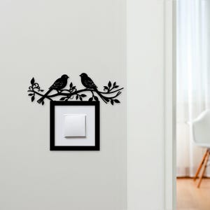 May include: Black metal wall art featuring two birds perched on a branch above a light switch. The birds and branch are a silhouette design. The light switch is white with a black frame, mounted on a white wall.