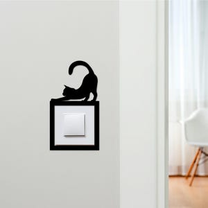 Könnte beinhalten: Schwarze Katzensilhouette, die sich über einem Lichtschalter streckt. Die Katze ist gewölbt, mit ausgestreckten Vorderpfoten und gebogenem Schwanz. Der Lichtschalter ist weiß, in einem schwarzen Rahmen, an einer weißen Wand montiert. Ein dekoratives Element für ein Zuhause.