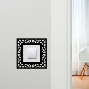 May include: A black decorative light switch cover with a geometric design. The cover surrounds a white light switch on a white wall. The design is square with a detailed pattern. The background is a white wall and a doorway.