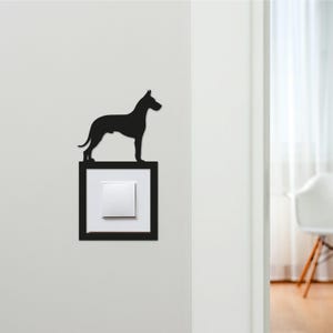 May include: A black silhouette of a dog stands atop a black framed light switch. The light switch is white and square. The dog is a sleek, stylized representation of a canine, likely a breed such as a Doberman or similar.