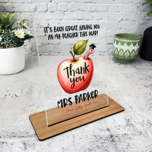 Puede incluir: Placa de agradecimiento para profesores de acrílico transparente con base de madera. La placa presenta una ilustración de una manzana con un gusano con gorro de graduación, y el texto "Thank you MRS PARKER From Lilly Smith". El texto superior dice "IT'S BEEN GREAT HAVING YOU AS MY TEACHER THIS YEAR!"
