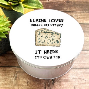May include: A round, silver tin with a white lid. The lid has the text "ELAINE LOVES CHEESE SO STINKY" and "IT NEEDS ITS OWN TIN", plus an illustration of blue cheese.