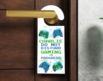 Könnte beinhalten: Weißer Türhänger mit einem pixelartigen Design in Grün und Blau, das einen Videospiel-Controller und den Text "Charlie Nicht stören Gaming in Progress" zeigt.