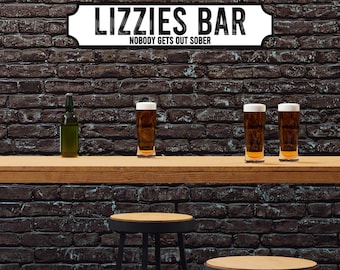 May include: A black and white sign with the text "Lizzie's Bar" and the tagline "Nobody gets out sober". The sign is hanging on a brick wall above a wooden bar counter with three glasses of beer and a bottle of beer. There are two bar stools in front of the counter.