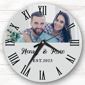 Op de afbeelding: Een witte ronde klok met Romeinse cijfers en een zwarte wijzer. De wijzerplaat van de klok heeft een foto van een stel en de tekst "Henry & Rose EST. 2023".