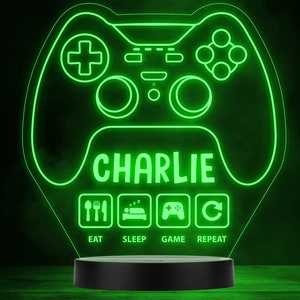 Könnte beinhalten: Eine grüne LED-Lampe in Form eines Gamecontrollers. Der Name "CHARLIE" wird in der Mitte angezeigt, darunter die Worte "EAT, SLEEP, GAME, REPEAT". Die Lampe steht auf einem schwarzen Sockel.