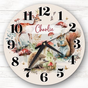 Op de afbeelding: Een crèmekleurige ronde klok met zwarte cijfers en wijzers. De klok heeft een aquarelontwerp van een hert, eekhoorn en egel in een bosomgeving. De naam "Charlie" staat in rood op de wijzerplaat van de klok.