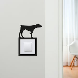 May include: A black silhouette of a dog standing on a light switch cover. The light switch cover is white with a black frame. The dog is a Pointer breed. The wall is white.