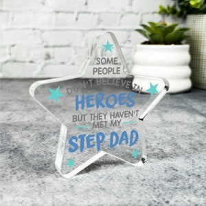 May include: A clear acrylic star-shaped sign with the text "Some people don't believe in heroes but they haven't met my step dad". The text is in blue and gray with small blue stars around the text.