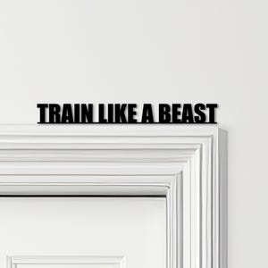 May include: A black metal sign displaying the words "TRAIN LIKE A BEAST" above a white door frame. The text is in a bold, sans-serif font, offering a clear and impactful message. The stark contrast of black against white enhances readability.