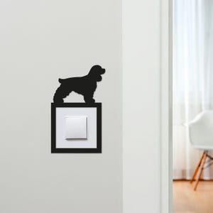 May include: A black silhouette of a dog, likely a Cocker Spaniel, perched above a light switch. The light switch is set in a black frame against a white wall. The dog is facing left, with its tail and ears visible.