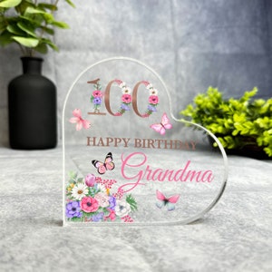 May include: A clear heart-shaped acrylic sign with a floral design and butterflies. The number 100 is in the center with a rose gold finish. The text "Happy Birthday Grandma" is below the number.