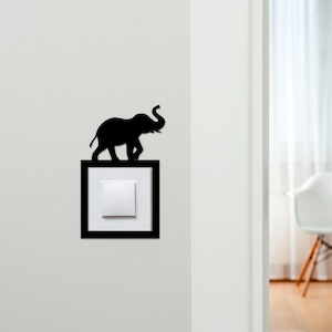 May include: A black elephant silhouette decorates a light switch cover. The elephant is positioned above a white light switch within a black rectangular frame, mounted on a white wall. The design adds a decorative touch to a functional item.