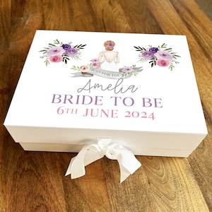 May include: White gift box with a purple and pink floral design and a bride illustration. The box has a white ribbon and the text "Just for you Amelia Bride to be 6th June 2024".