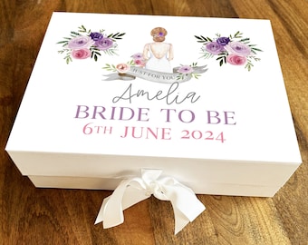 Puede incluir: Caja de regalo blanca con un diseño floral morado y rosa y una ilustración de novia. La caja tiene una cinta blanca y el texto "Just for you Amelia Bride to be 6th June 2024".