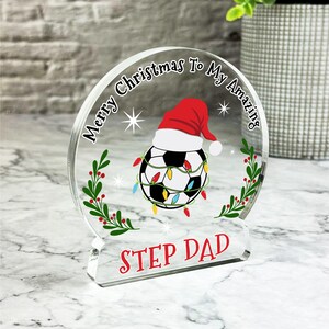 May include: A clear acrylic Christmas ornament with a soccer ball wearing a Santa hat and surrounded by Christmas lights. The ornament reads "Merry Christmas To My Amazing Step Dad".